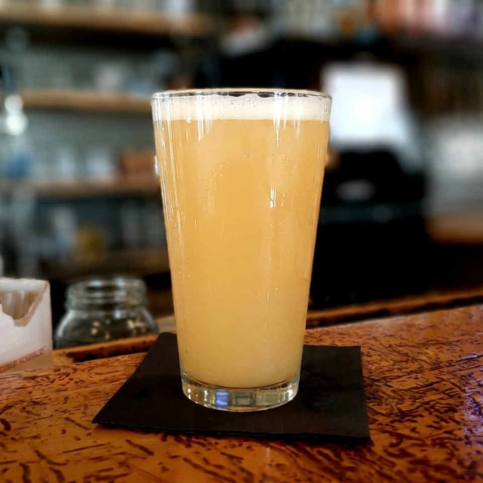 Pint glass of hazy golden beer with a foamy head on a black coaster atop a wooden bar.